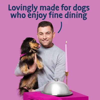Text reads: Lovingly made for dogs who enjoy fine dining. Image shows a person holding a dog next to a table with a covered dish and a lit candle.