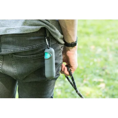 Pojemnik na woreczki na odchody przypięty do szlufki spodni za pomocą karabińczyka, widoczny fragment smyczy w dłoni osoby.