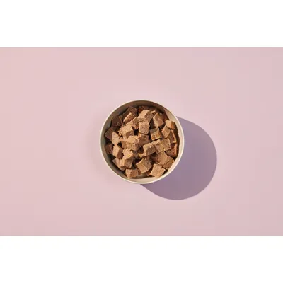 Bowl filled with small, cubed pieces of brown pet food or treats, shown from above on a plain background.
