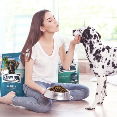 Happy Dog Karibik torrfoder i skål, dalmatiner matas. Förpackning i bakgrunden visar produkten Happy Dog Sensible Medium Adult.