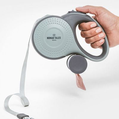 Mano che tiene un guinzaglio grigio per cani con dispenser integrato per sacchetti igienici. Cinturino grigio e moschettone in metallo visibili.