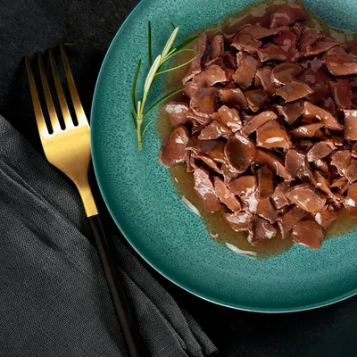 Närbild av en grön tallrik med saftiga köttbitar i sås, dekorerad med en rosmarinkvist. Guldgaffel och svart tygservett bredvid.