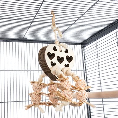 Zawieszana zabawka z drewna i plecionego materiału z motywami serc, kulkami i sznurkami, zamocowana w klatce dla ptaków.