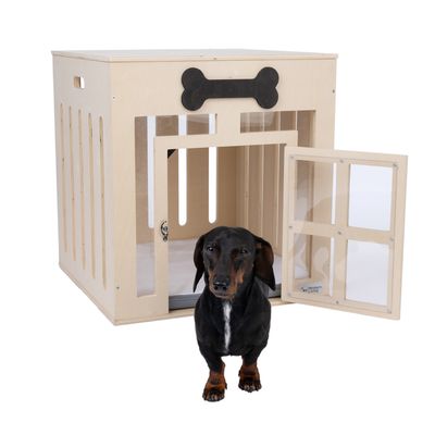 Niche ou cage pour chien en bois clair avec porte transparente ouverte, motif os noir sur le dessus, un teckel noir et feu debout devant l’entrée. Marque Modern Living visible sur la porte.