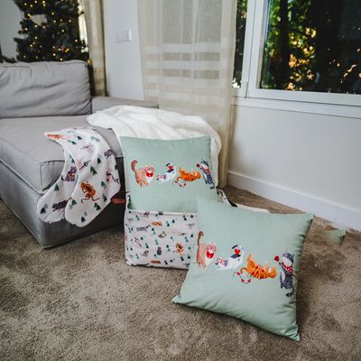 Prydnadskudde och filt med kattmotiv i juldesign, arrangerade på en soffa och matta.