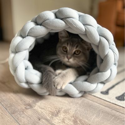Gato gris tumbado en un túnel trenzado de tela gris claro y suave sobre suelo de madera.