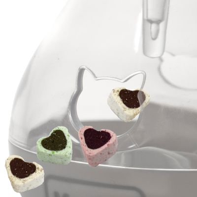 Friandises en forme de cœur, couleurs vert, rose et blanc, devant un distributeur transparent avec ouverture en forme de tête de chat. Aucun texte visible.
