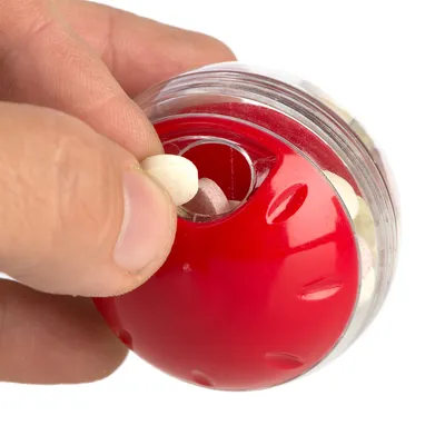 Main insérant une friandise ronde dans une boule en plastique rouge et transparent remplie de petites friandises. Matériaux visibles : plastique.