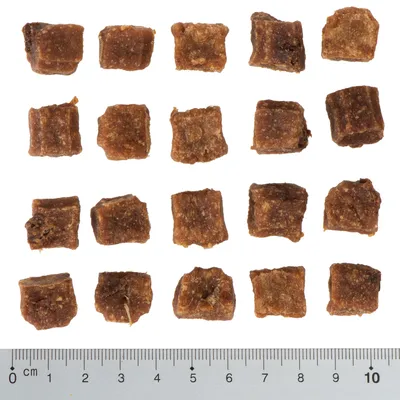 Morceaux de friandises pour animaux en forme de cubes, disposés en rangées sur fond blanc avec règle en centimètres montrant une taille d’environ 1,5 cm par cube.