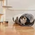 Katze sitzt in einem grauen und weißen Filztunnel auf Holzboden. Im Hintergrund ein Kratzbaum und eine Metallfutterschale sichtbar.