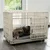 Hund liegt in einem beigen Metallkäfig mit Gitterwänden und einer grauen Matte auf dem Boden. Käfig hat eine Tür und Rollen für Mobilität.