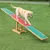 Perro sobre balancín de agilidad para perros, estructura de madera con superficie antideslizante verde y roja, colocado en césped al aire libre.