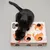 Schwarze Katze spielt mit Tiaki Spielbox aus Pappe, Löcher und orangefarbene Katzenmotive sichtbar, gelb-rosa Ball im Inneren.