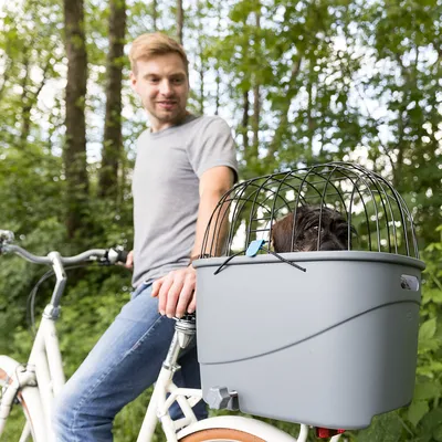 Panier gris en plastique avec grille métallique pour animal, fixé à l’arrière d’un vélo blanc, petit chien noir visible à l’intérieur du panier.