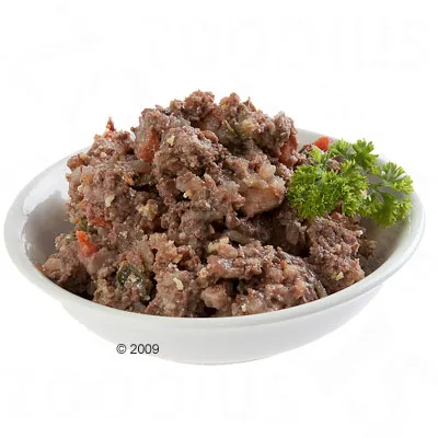 Comida húmeda para mascotas servida en un cuenco blanco, mezcla de carne picada con trozos visibles de verduras y una ramita de perejil como adorno.