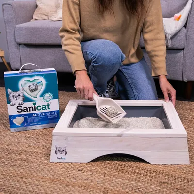 Litière pour chat Sanicat Active White, boîte visible à gauche, personne utilisant une pelle pour retirer un agglomérat dans un bac à litière en bois clair.