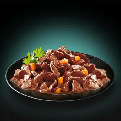 Kawałki mokrej karmy dla zwierząt na czarnym talerzu, widoczne brązowe kawałki mięsa w sosie z dodatkiem pomarańczowych warzyw i zielona natka pietruszki.