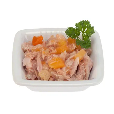 Влажный корм для животных в белой керамической миске, кусочки мяса с оранжевыми и желтыми добавками, украшено веточкой петрушки.