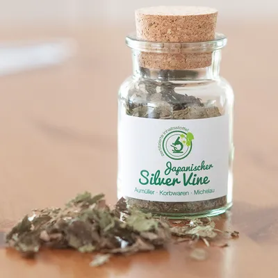 Skleněná láhev s korkovým uzávěrem, naplněná sušenými listy. Štítek: Japanischer Silver Vine, certifikované složky, Aumüller Korbwaren Michelau.