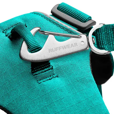 Particolare di una pettorina in tessuto turchese con gancio metallico inciso 'RUFFWEAR' e fibbia regolabile visibili.