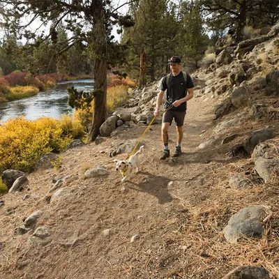 Person walking a small white and brown dog on a yellow lead along a rocky forest path beside a river, both wearing outdoor gear. Trees and bushes visible in the background.