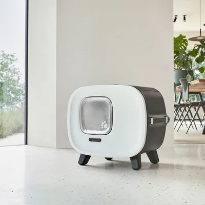 Modern cat house with white front, transparent door with paw motif, and black sides. Stands on four legs in a bright room.