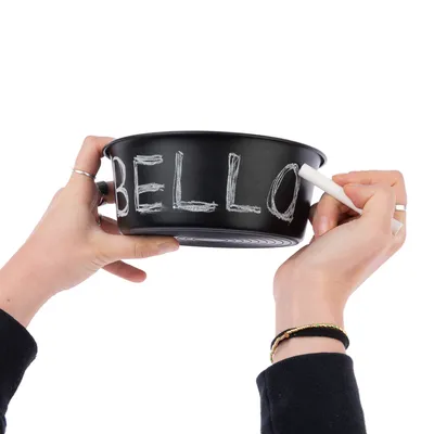 Musta ruokakuppi liitutaulupinnalla, jossa lukee 'Bello'. Kaksi kättä pitää kuppia ja kirjoittaa siihen valkoisella liidulla.