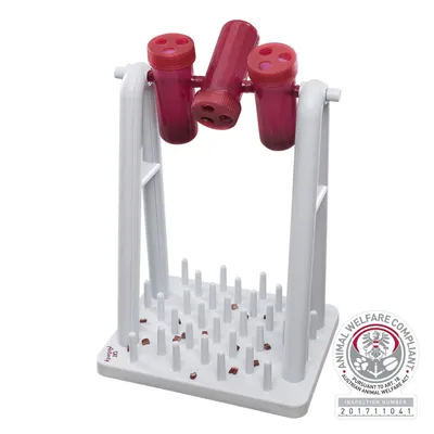 Hvid og rød aktivitetslegetøj med tre roterende beholdere med huller, stående på en base med lodrette plastikpinde. Mærke: Animal Welfare Compliant, inspection number 201711041.