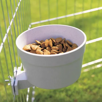Gamelle en plastique fixée à une grille, remplie de croquettes marron. Système de fixation métallique visible sur le côté gauche du bol.