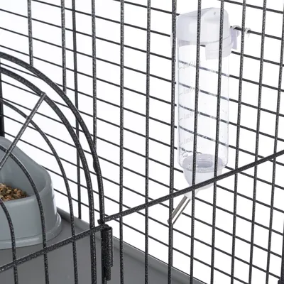 Sticlă de apă din plastic transparent cu ieșire metalică, atașată de o cușcă cu grilaj negru. Lângă sticlă, un bol gri cu semințe.