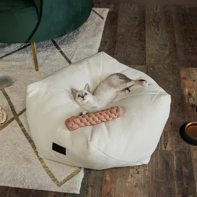 Cama para gatos de tela blanca con juguete trenzado rosa, gato tumbado relajado encima. Suelo de madera y alfombra.
