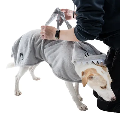 Dog wearing grey fleece coat with shark design. Person holding coat by carrying handle. 'Trixie' logo visible on side of coat.