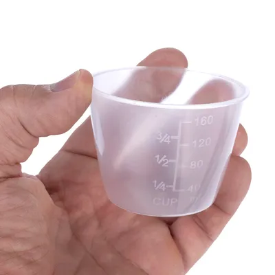 Målebæger af transparent plast med skala i milliliter og cups, maksimal kapacitet 160 ml. Bægeret holdes af en hånd.