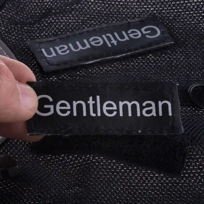 Sort etiket med hvidt skrift 'Gentleman', holdt af en hånd. Baggrunden viser et andet etiket og netmateriale.