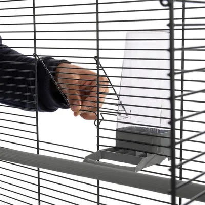 Hand opening a cage door next to a transparent plastic feeder attached to the metal bars of the cage.