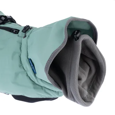 Vue rapprochée d’un manteau pour chien vert clair avec doublure polaire grise, capuche ajustable et fermeture éclair visible. Étiquette bleue avec texte partiellement lisible.