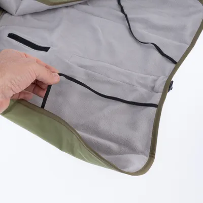 Vue intérieure d’un manteau pour animal en tissu vert et gris, doublure polaire, poche zippée visible et élastiques noirs tenus par une main.