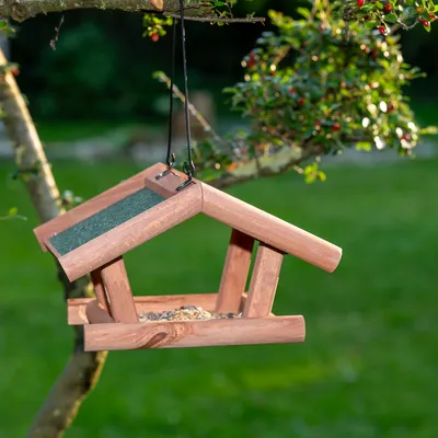 Casita para pájaros colgante de madera con techo verde, llena de alimento para aves, fijada a una rama en el jardín.