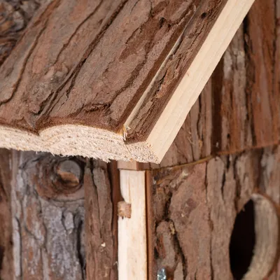 Närbild av ett fågelhus i naturträ med barkbeklädnad och rund ingång. Synlig trästruktur och skruvar för montering.