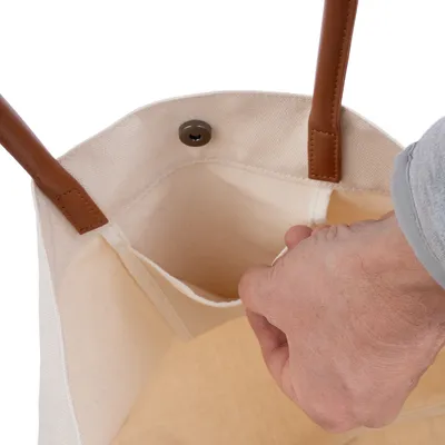 Bolsa de tela beige con asas marrones, bolsillo interior y cierre de botón a presión. Mano accediendo al bolsillo.
