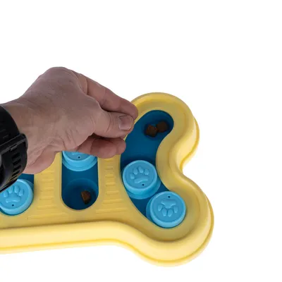 Main plaçant des croquettes dans un jouet d’intelligence pour chien en forme d’os, plastique jaune et bleu avec compartiments à couvercles bleus décorés de pattes.