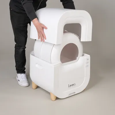 Person demonstrating assembly of a white kooa cat toilet with removable lid and round inner chamber. Wooden legs and controls visible.