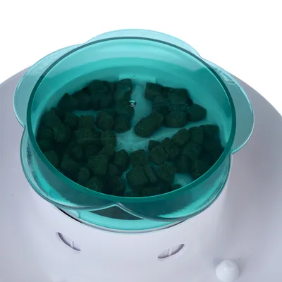 Grünes Trockenfutter in einem transparenten, grünen Behälter mit weisser Basis. Sichtbare Öffnungen an der Basis des Behälters.