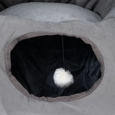 Grey pet bed with cave entrance and hanging white plush ball. Interior made of black fabric, top lying surface made of soft grey material.