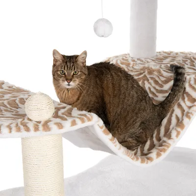 Smeđa tigrasta mačka sjedi na visećoj ležaljci od bež i bijelog pliša, dio grebalice s užetom i visećom bijelom lopticom vidljiv u kadru.