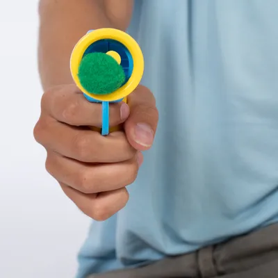 Hand hält gelb-blauen Ballwerfer mit grünem Filzball, geeignet für interaktives Spielen.