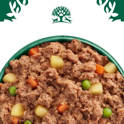 Nærbillede af vådfoder med kødstykker, gulerødder, ærter og kartoffeltern i en grøn skål. Logo med træ og dyr øverst synligt.