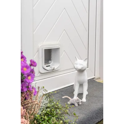 Katzenklappe in weißer Tür, daneben eine weiße Katzenfigur und ein graues Stoffspielzeug in Mäuseform auf einer Matte. Katzenklappe in weißer Tür, daneben eine weiße Katzenfigur und ein graues Stoffspielzeug in Mäuseform auf einer Matte.