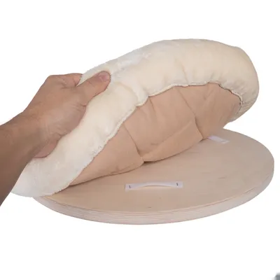 Coussin rond beige à bordure crème en tissu et fausse fourrure, soulevé par une main, posé sur une base circulaire en bois avec bandes auto-agrippantes visibles.
