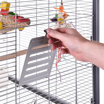 Mano colocando una bandeja metálica en la puerta de una jaula para pájaros, con juguetes colgantes y accesorios visibles en el interior.